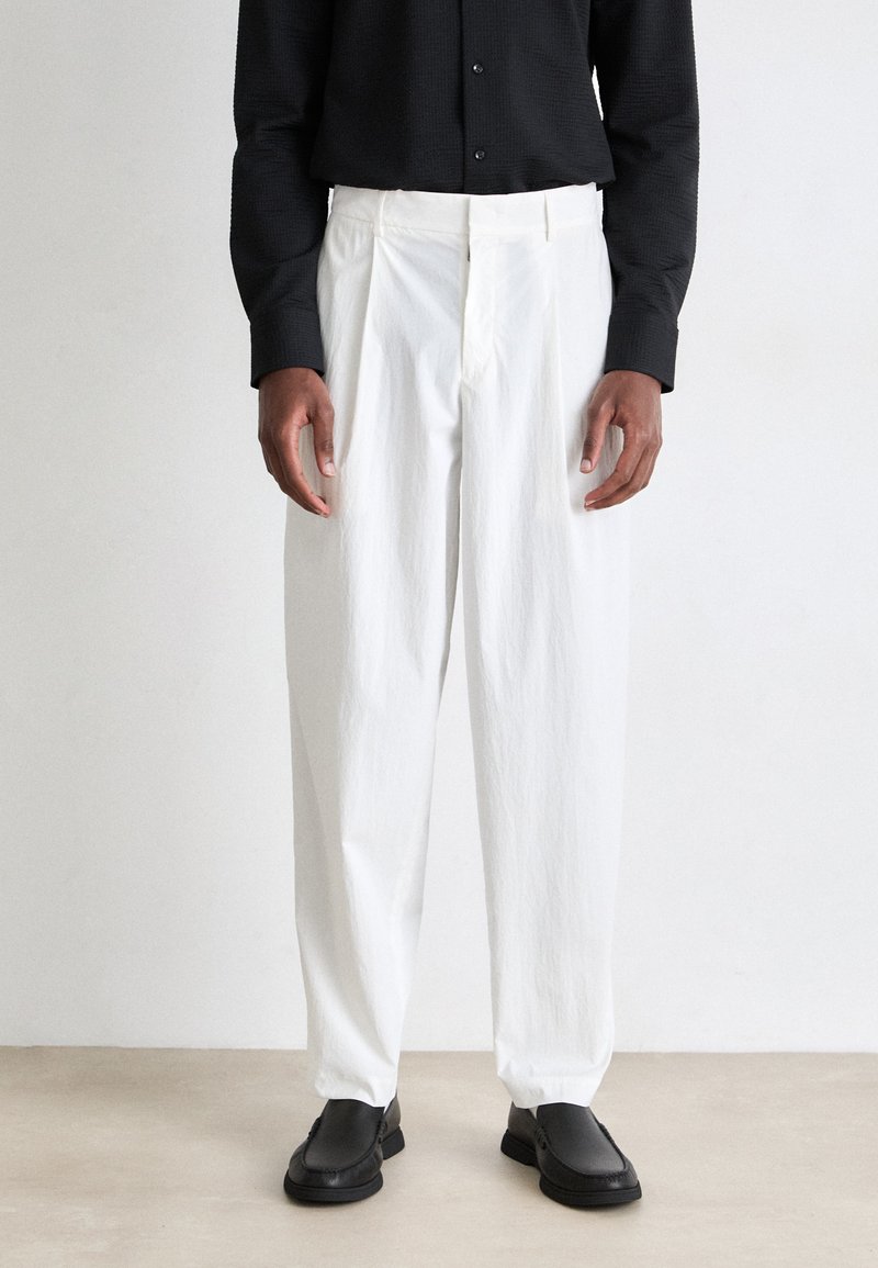 White pleated trousers made of textured fabric, featuring a relaxed fit and paired with black shoes, worn with a black button-up shirt.
