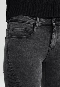 Pantalones vaqueros de mezclilla negra con un acabado deslavado. Las características incluyen un bolsillo frontal, detalles de costura y un ajuste slim. El material es texturizado y ligeramente elástico.