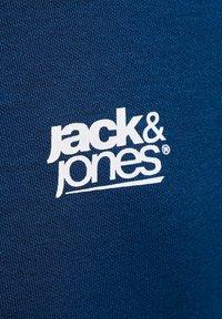 Λευκό λογότυπο "jack & jones" τυπωμένο σε υφασμάτινη υφή μπλε χρώματος, κεντραρισμένο με μια ελαφριά υπογράμμιση κάτω από το "jones".