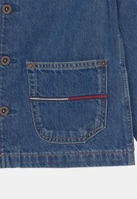 Jeansjacka med ficka, bruna knappar, beige sömmar och en liten randig detalj i rött, vitt och blått vid fickans kant.