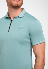Šviesiai mėlynas polo marškinėliai su užtrauktuku ant apykaklės, trumpomis rankovėmis, tekstūruota medžiaga ir mažas logotipas ant krūtinės. Akcentuoti pilkų detalių.