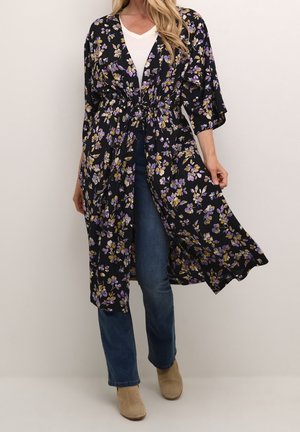 Mujer con un cárdigan negro estilo kimono con flores sobre una camiseta blanca, vaqueros azules y botines beige sobre un fondo liso.