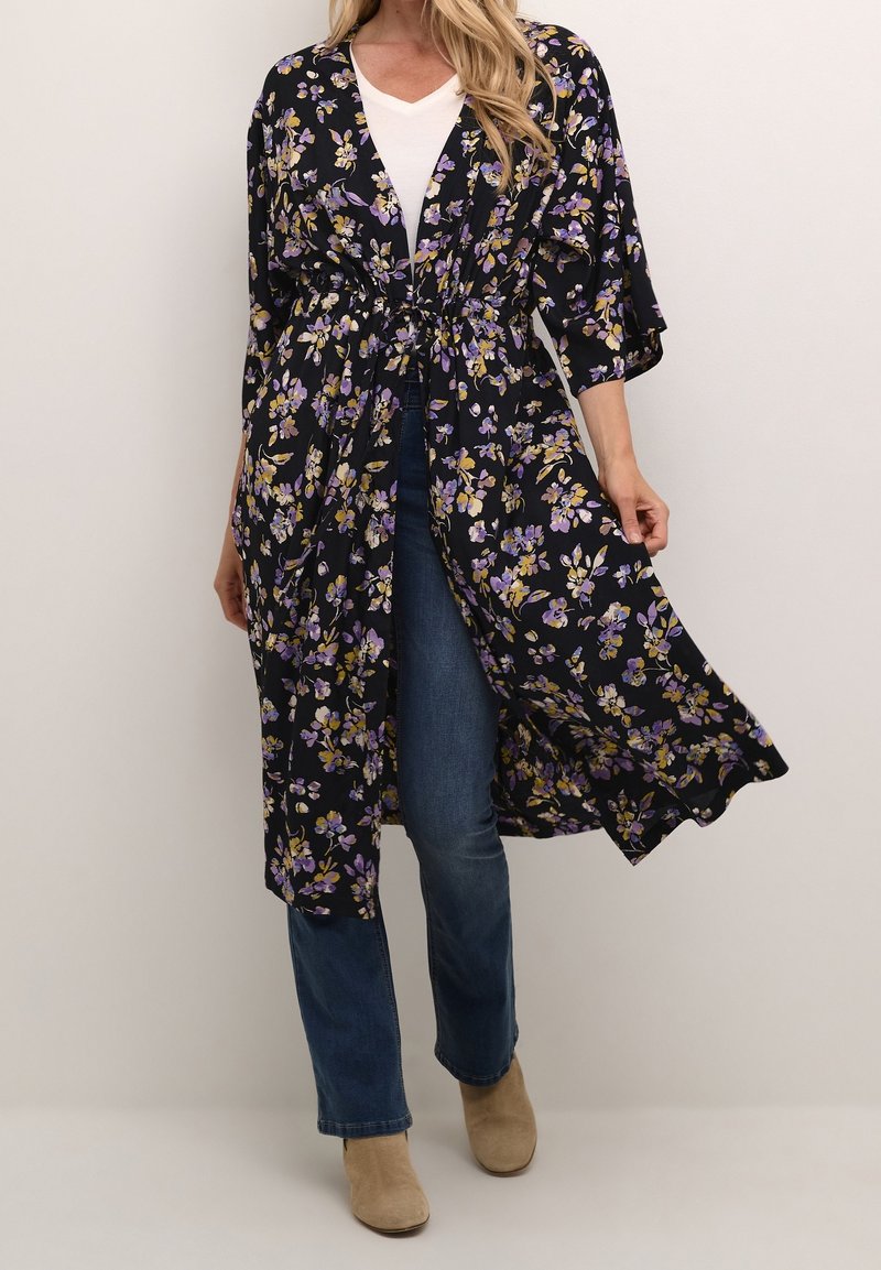 Femme portant un cardigan noir style kimono à motifs floraux sur un top blanc, un jean bleu et des bottines beiges sur un fond uni.