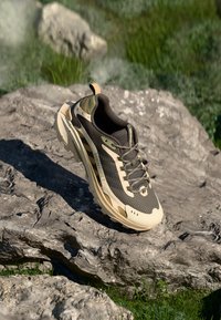 Trail shoe with a beige and olive color scheme, mesh and synthetic upper, textured sole, and prominent lacing system on a rocky surface.
