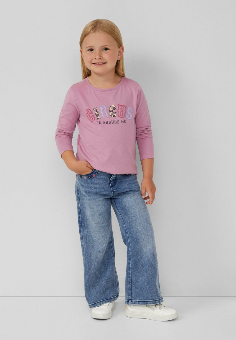 Helles rosa Langarmhemd mit "CIRCUS IS AROUND ME" in bunten Buchstaben bedruckt. Blaue weite Jeans mit dekorativen Knöpfen. Weiße Turnschuhe.