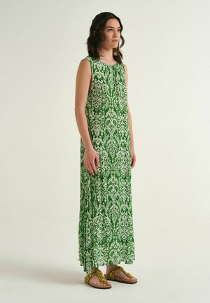 Donna con capelli scuri e mossi che indossa un abito lungo senza maniche verde e bianco a fantasia e sandali dorati con perline, in piedi davanti a uno sfondo semplice.