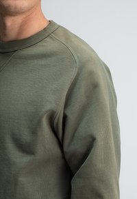 Olijfgroene sweatshirt met een ronde halslijn, gemaakt van een zachte, gestructureerde stof en raglanmouwen. Gladde afwerking met minimale naden.