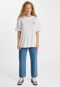 Biała bawełniana koszulka z luźnym krojem i krótkimi rękawami, z zielonym nadrukiem na lewej piersi. Połączona z niebieskimi dżinsami o prostych nogawkach.