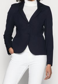 Blazer blu navy realizzato in tessuto morbido, caratterizzato da una vestibilità su misura, rever a punta, due bottoni e tasche frontali. Abbinato a un top bianco.
