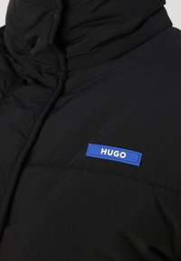 Fekete párnázott dzseki magas gallérral. Kék, téglalap alakú címke található rajta, amelyen a "HUGO" felirat olvasható. Az anyag sima, matt textúrájú.