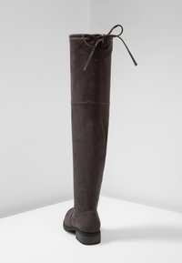 Bota de ante marrón oscuro, alta, con un tacón bajo y un lazo atado en la parte trasera superior, posicionada sobre una superficie blanca frente a un fondo gris claro.