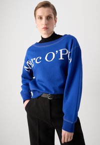 Zils džemperis ar baltu tekstu, kas lasīts "Marc O’Polo", ar plašu kakla izgriezumu, ribotām piedurknēm un kombinēts ar melnām biksēm.