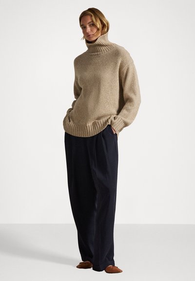Ženska, ki stoji z rokami v žepih, oblečena v bež pulover s turtleneck ovratnikom, ohlapne temno modre hlače in rjave nizke čevlje, pred enobarvnimi ozadjem.