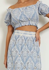 Blauwe off-the-shoulder crop top met pofmouwen, voorzien van witte kant borduursels en oogjesdetails. Gepaarde met een bijpassende rok.