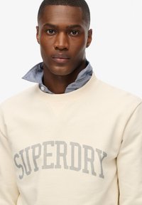 Sweatshirt crème à col rond, arborant un logo gris "SUPERDRY" bien visible. Chemise bleue à col visible en dessous.