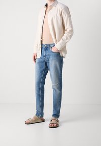 Avslappnad outfit som består av en ljusbeige skjorta med knappar, en krämfärgad ribbad linne, ljusblå tapered jeans med slitna områden och beige sandaler.