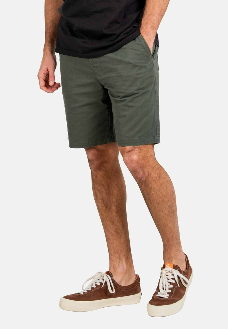 Pantalones cortos verde oliva, hechos de una mezcla de algodón, con bolsillos laterales y un corte relajado. Combinados con zapatillas de ante marrón y cordones blancos.