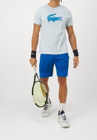 Light blue t-shirt featuring a blue crocodile graphic, paired with blue shorts. The model holds a tennis racket, wearing white athletic shoes.