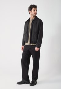 Chaqueta ligera negra con dos bolsillos frontales, camiseta beige debajo, pantalones negros y zapatos negros, con un fondo neutro.