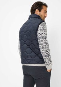 Marineblaue gesteppte Weste mit hohem Kragen, kombiniert mit einem gemusterten Pullover mit geometrischen Designs in Marineblau und Creme. Dunkle Hosen sind sichtbar.