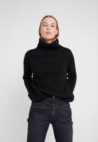 Black ribbed turtleneck sweater with a relaxed fit, featuring raglan sleeves and a high collar. Paired with dark blue denim jeans.