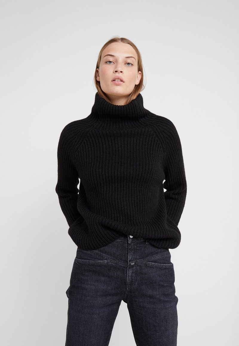 Black ribbed turtleneck sweater with a relaxed fit, featuring raglan sleeves and a high collar. Paired with dark blue denim jeans.