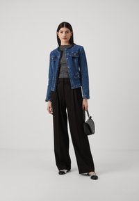 Spijkerjacket met knoopsluitingen en twee voorkanten pockets, gecombineerd met een textuurgrijze top en zwarte wijde broek. Zwarte handtas en schoenen.