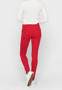 Jeans ajustados rojos con un ajuste ceñido y dos bolsillos traseros. Combinados con un suéter blanco de canalé y zapatillas blancas. Textura suave, sin patrones visibles.
