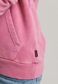 Felpa rosa con tasca a marsupio, orlo e polsini a costine. Presenta un piccolo etichetta nera del marchio sul lato.