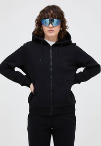 Zwarte zip-up hoodie met kangoeroezakken, verstelbare trekkoordcapuchon en geribbelde boorden. Draagt met grote, spiegelende zonnebril.