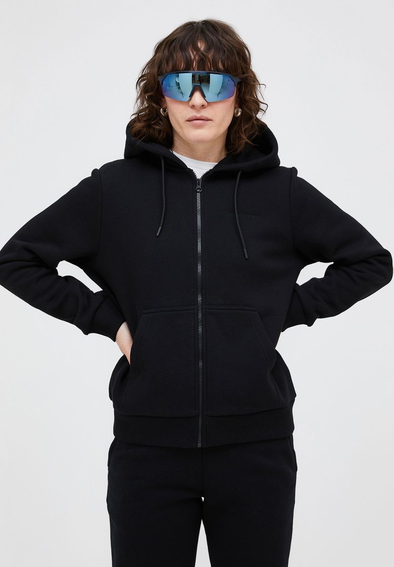 Zwarte zip-up hoodie met kangoeroezakken, verstelbare trekkoordcapuchon en geribbelde boorden. Draagt met grote, spiegelende zonnebril.