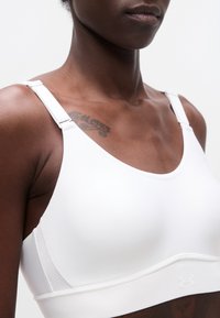 Κοντινό πλάνο ενός ατόμου που φοράει λευκό αθλητικό σουτιέν με ρυθμιζόμενες τιράντες και ορατό τατουάζ στο επάνω μέρος του στήθους.