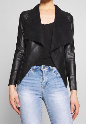 Mujer que lleva una chaqueta de cuero negra abierta al frente con solapas anchas sobre una blusa negra y jeans de talle alto de color azul claro.