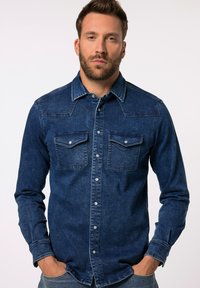Mørkeblå denimskjorte med knaplukning foran, to brystlommer og lange ærmer med knapmanchetter. Slidte teksturer er tydelige.