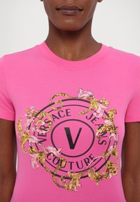 Rozā kokvilnas t-krekls ar apļveida grafiku, kurā ir uzraksts "Versace Jeans Couture", izsmalcināti zeltaini un rozā ziedu motīvi, kā arī centrā melns "V".