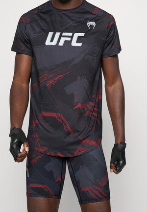 Männlicher Athlet trägt schwarzes und rotes UFC-Sportshirt und Shorts mit schwarzen fingerlosen Handschuhen, stehend vor einfarbigem Hintergrund.