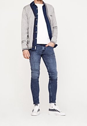 Muž oblečený v šedém kardiganu přes tmavomodrou košili na knoflíky a bílé tričko, v modrých přiléhavých džínách a bílých teniskách, stojící s rukou v kapse.