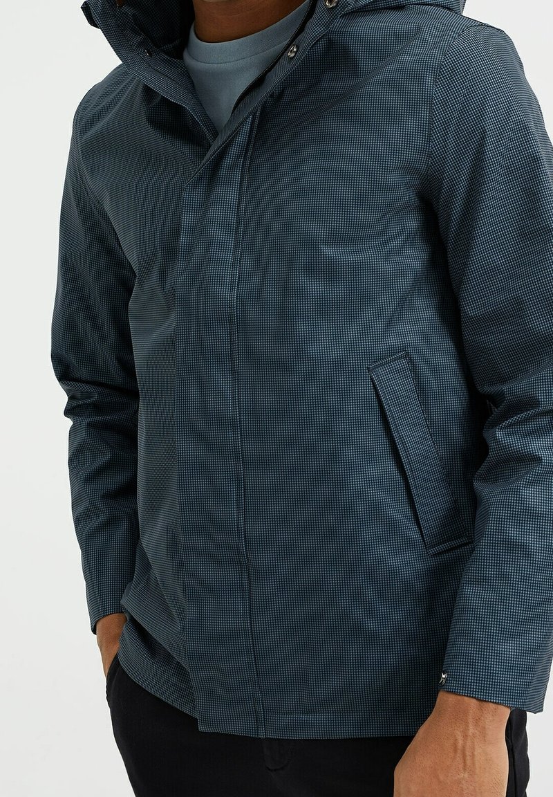 Homme portant une veste à capuche à carreaux bleus avec des poches avant, debout avec une main dans la poche et une chemise bleu clair en dessous.