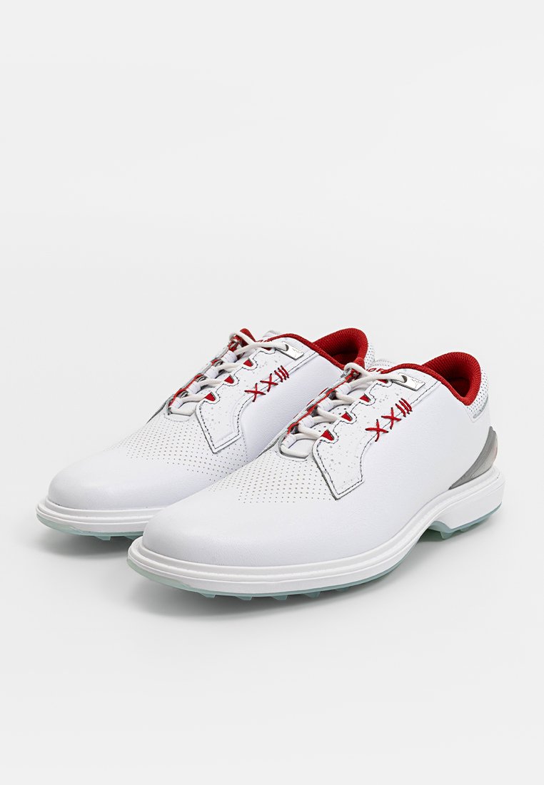Des chaussures de golf blanches avec une tige en cuir lisse, des accents rouges sur la languette et les lacets, des détails perforés et une semelle courbée gris clair.