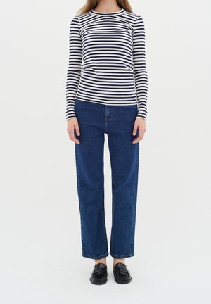 Osoba ubrana w czarno-białą bluzkę w paski z długim rękawem, proste niebieskie dżinsy oraz czarne buty wsuwane, stojąca na tle jednolitego tła.