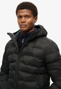 Chaqueta de plumas negra con capucha, con un diseño acolchado y cremallera en la parte frontal. Usada sobre un suéter de punto azul marino con detalles de botones.