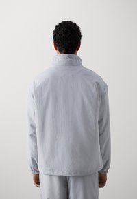 Chaqueta gris claro con un cuello alto, puños elásticos y un corte relajado; presenta una textura suave y mate y un diseño minimalista.