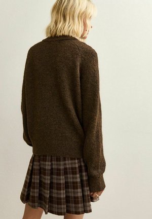 Un pull en laine marron associé à une jupe plissée à carreaux marron avec des accents blancs. La texture semble douce et confortable, avec une coupe décontractée.