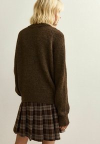 Brązowy sweter wełniany zestawiony z brązową spódnicą plisowaną w kraciasty wzór z białymi akcentami. Tekstura wydaje się miękka i przytulna, z luźnym krojem.