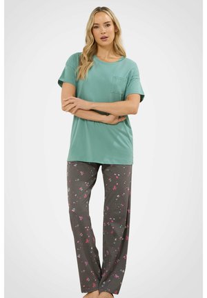 Kobieta o długich blond włosach, ubrana w zieloną koszulkę z krótkim rękawem oraz ciemne spodnie do spania w różowe i niebieskie wzory serc oraz kokardek.