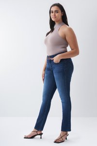 Top sin mangas beige con cuello alto, combinado con jeans ajustados de color azul oscuro. Sandalias negras de tacón con tiras delgadas completan el look.