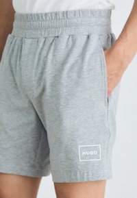 Grå idrettsshorts med elastisk midjebånd, sidelommer og en hvit rektangulær logo med "HUGO" nederst til venstre. Myk stofftekstur.
