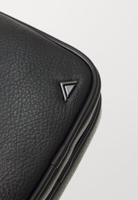Angolo di una borsa in pelle nera testurizzata con un accento metallico triangolare e una cucitura liscia e curvata lungo il bordo.