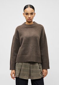 Γυναίκα με μαλλιά χτενισμένα προς τα πίσω, που φοράει καφέ ριμπ πουλόβερ πάνω από καρό πλισέ φούστα και σκούρο παντελόνι, όρθια μπροστά σε απλό φόντο.