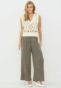 Elefántcsont színű, ujjatlan horgolt felső virágmintás kivágásokkal, széles szárú, texturált szürke nadrággal és fehér, hegyes orrú magassarkúval párosítva.